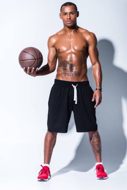full length view of athletic shirtless african american man with basketball ball looking at camera on grey  clipart