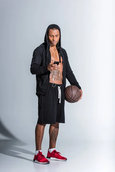 美国黑人男子篮球戴着耳机和使用智能手机灰色 — 图库照片