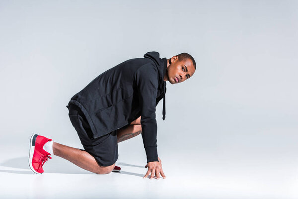 side view of young african american sportsman in hoodie ready to run and looking at camera on grey
