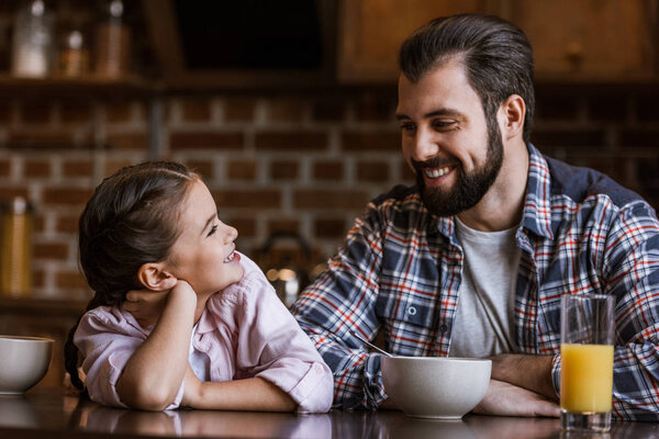 happy father with daughter sitting at table with juice and bowls and looking at each other at kitchen