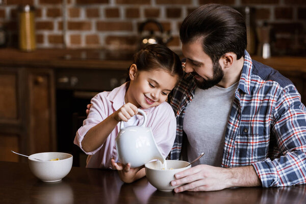 father and daughter pouring milk in bowl with snacks at kitchen