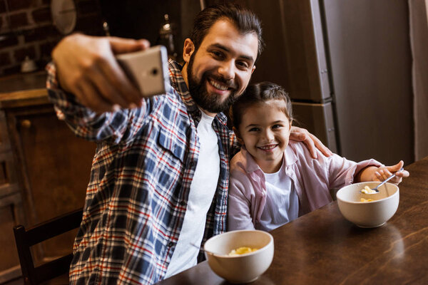 happy father with daughter sitting at table with snacks in bowls and takes selfie at kitchen