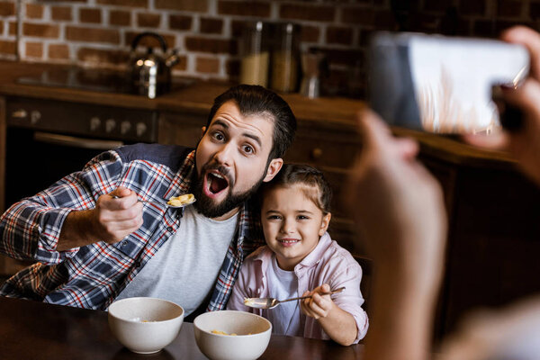 woman takes photo of father with daughter sitting at table and eating snacks from bowls at kitchen