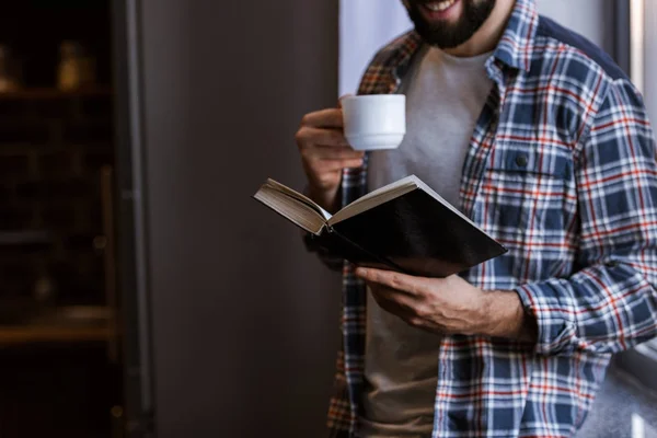 与咖啡杯站在窗边看书的欢快男子的裁剪图像 — 图库照片