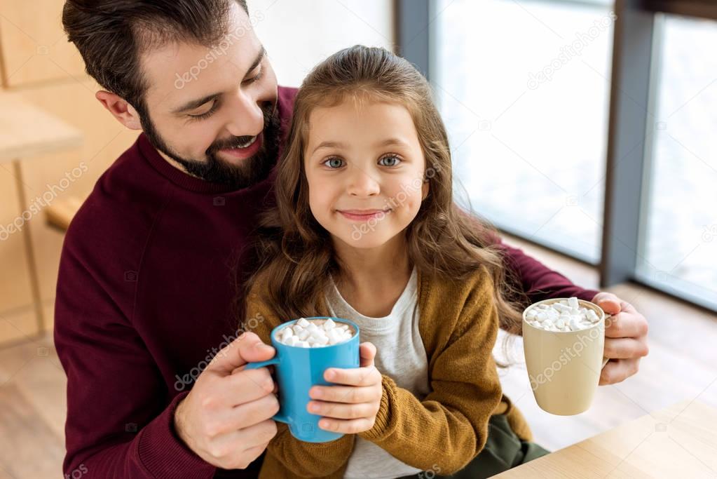 adorable daughter sitting on father knees and holding cup of cacao with marshmallow and looking at camera 