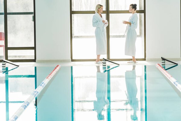beautiful young women standing next to swimming pool at spa center and talking with coffee cups in hands