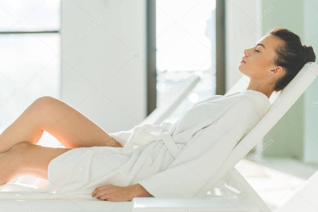 side view of beautiful young woman in bathrobe lying on sunbed at spa