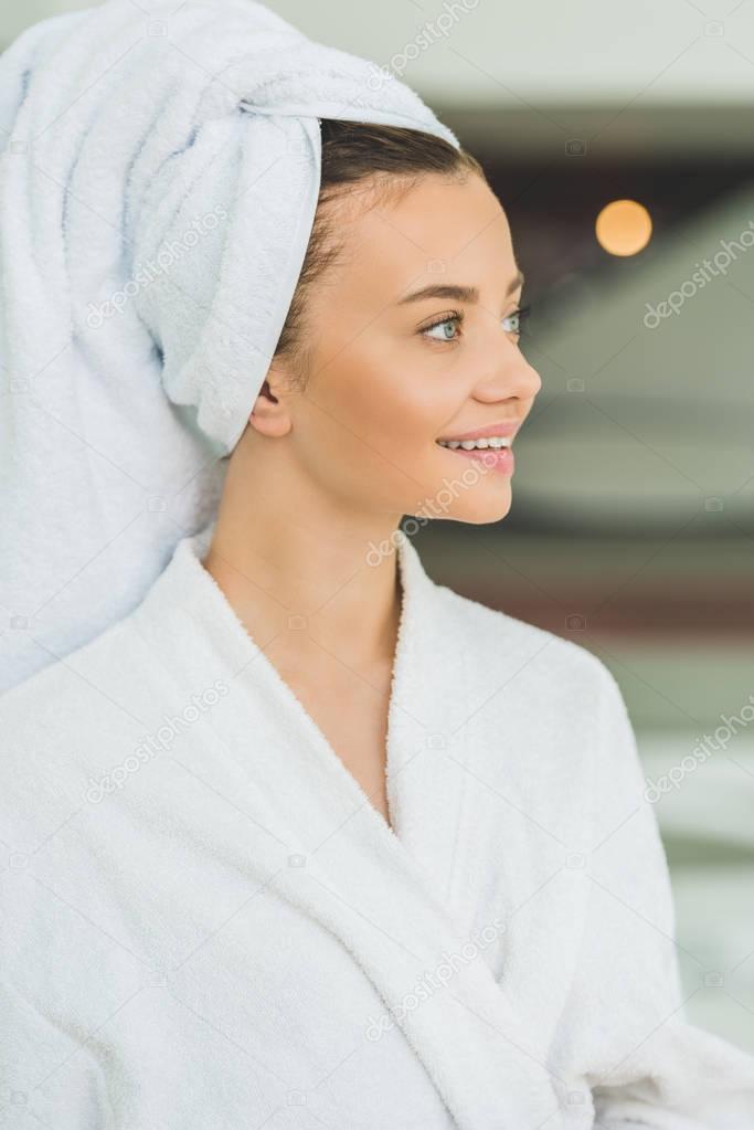smiling young woman in bathrobe and towel on head at spa salon