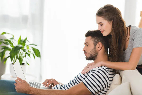 年轻女子拥抱她的男朋友 而他在家里的笔记本电脑的侧视图 — 图库照片