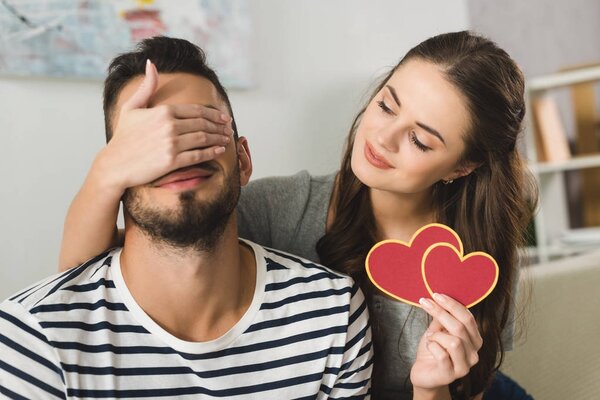 Молодая женщина закрывает глаза бойфренда рукой и держит открытку на день Святого Валентина
