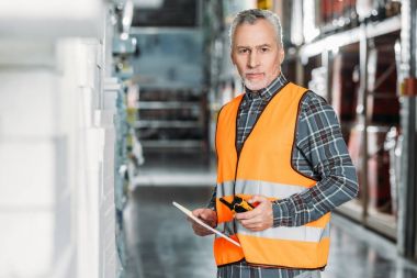 senior worker in safety vest using digital tablet and walkie talkie in storehouse clipart