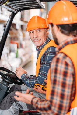 Emanet yelek ve kask forklift makine içinde oturan ve depolama alanındaki meslektaşı ile konuşmak kıdemli işçi