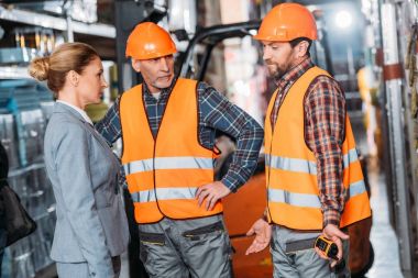 male workers in safety vests and helmets talking with inspector in storehouse clipart