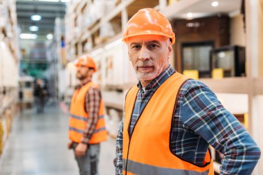 senior worker in safety vest and helmet, coworker standing behind in storage clipart