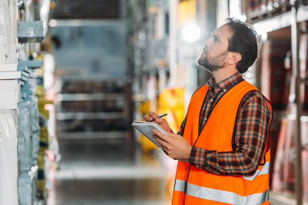 male worker in safety vest writing in notepad in storehouse