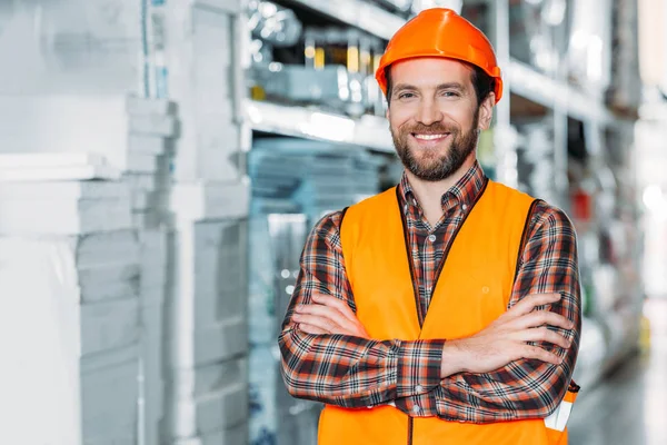 smiling worker in helmet and safety vest with crossed arms in warehouse