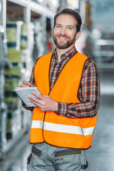 smiling worker in safety vest writing in notepad in storehouse