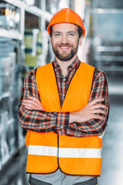 cheerful worker in helmet and safety vest with crossed arms in warehouse
