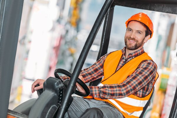 male worker in safety vest and helmet sitting in forklift machine in storage