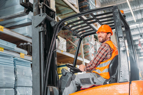 male worker in safety vest and helmet sitting in forklift machine in storage