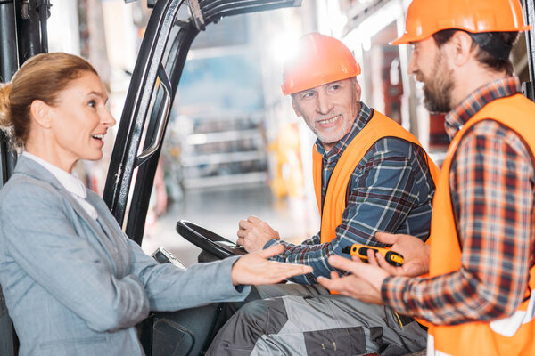 senior worker in safety vest and helmet sitting in forklift machine and talking with colleagues in storage