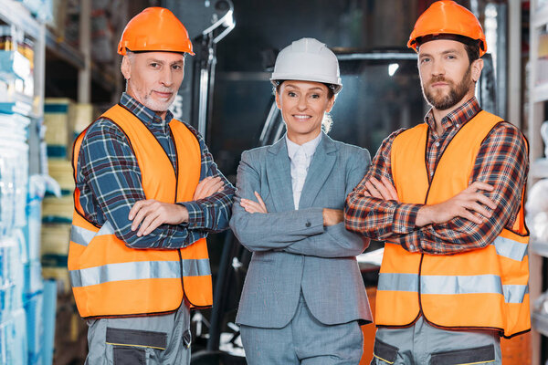 workers in helmets and inspector in suit posing with crossed arms in storage