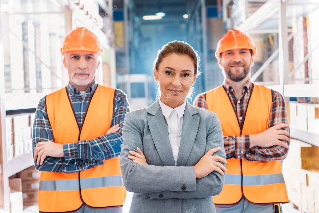 workers in helmets and inspector in suit posing with crossed arms in storage