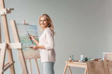 mature female artist smiling at camera while painting on easel in art studio clipart