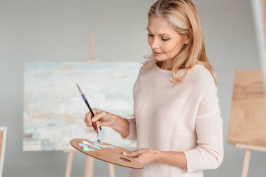 mature woman holding palette and paintbrush in art studio clipart