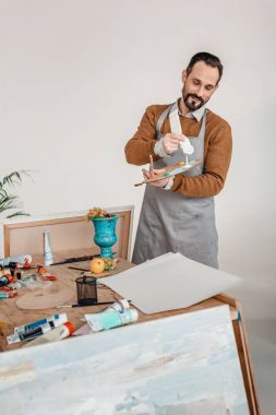 smiling mature male artist in apron holding palette in art studio clipart