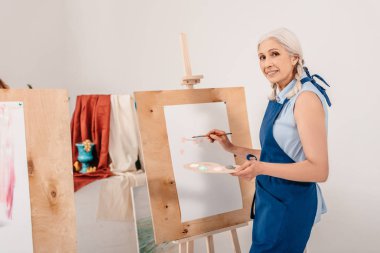 beautiful senior woman smiling at camera while painting on easel in art studio clipart