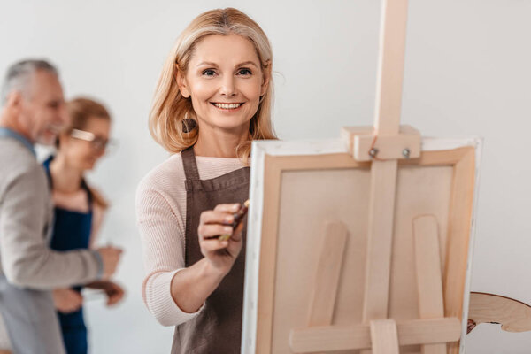 beautiful mature woman smiling at camera while painting on easel at art class 