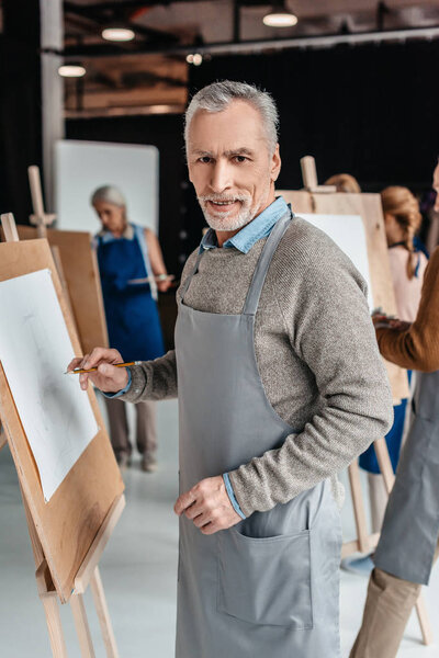 senior man smiling at camera while standing near easel at art class