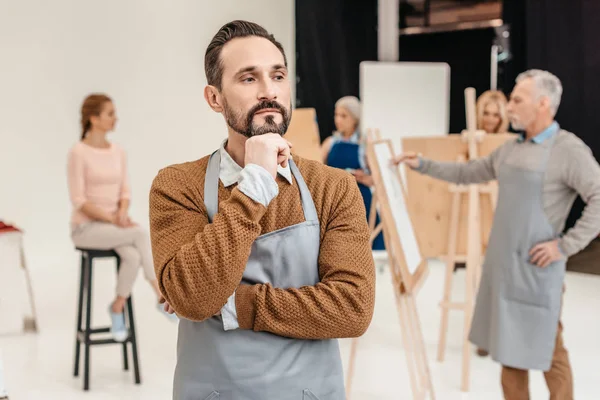 Pensive Bearded Artist Apron Looking Away While Adult Students Painting — Stock Photo, Image
