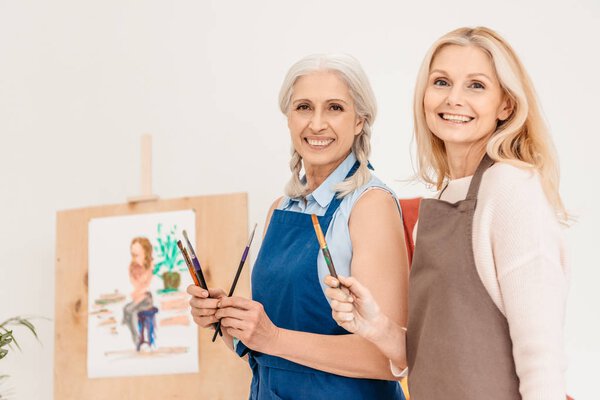 cheerful senior women smiling at camera and holding paint brushes at art class