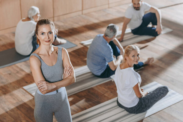 высокий угол обзора женщины-инструктора, смотрящей в камеру, в то время как пожилые люди занимаются на ковриках для йоги
