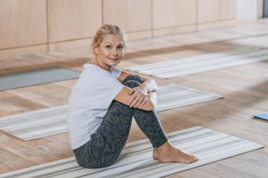 Yoga mat üzerinde oturan ve eğitim sınıf kameraya gülümseyen güzel Olgun kadın