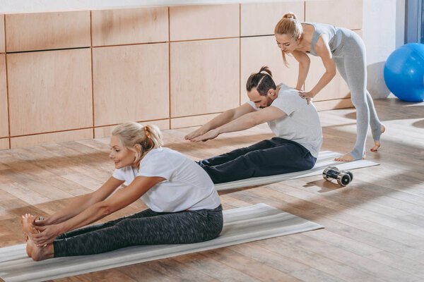 инструктор помогает взрослым людям растягиваться на ковриках для йоги на занятиях
