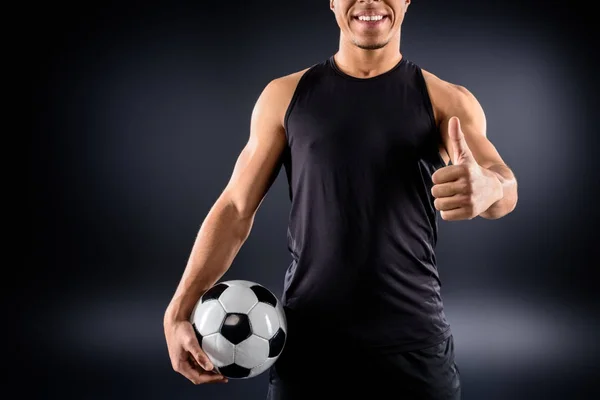 handsome african american soccer player with ball showing thumb up on black