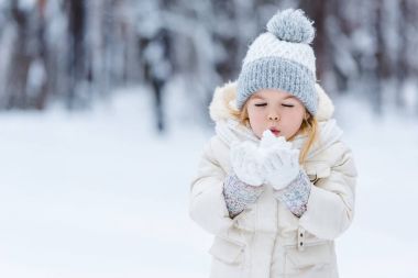 portrait of adorable kid blowing onto snow ball in hands in winter park clipart