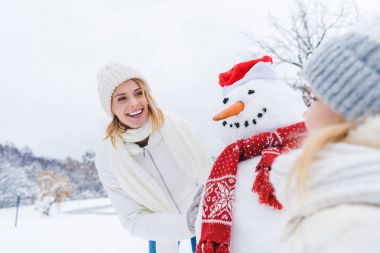 happy mother and daughter standing near snowman together in winter forest clipart