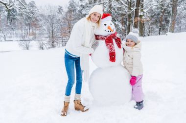 beautiful happy mother and daughter smiling at camera while standing with snowman in winter park clipart