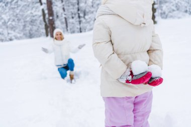 küçük kızı mutlu anne arkasında kış parkta diz çökmüş ve kartopu tutarak atış kırpılmış