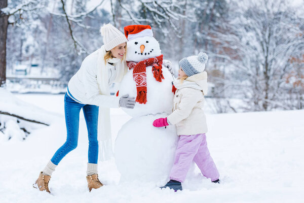 happy mother and daughter making snowman together in winter forest