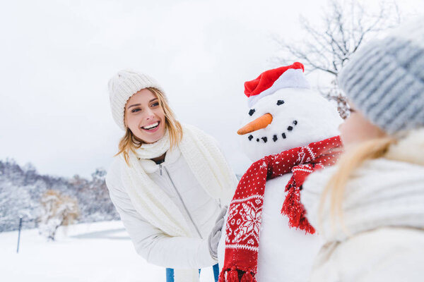 happy mother and daughter standing near snowman together in winter forest
