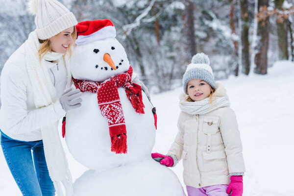 beautiful happy mother and daughter standing with snowman in winter park