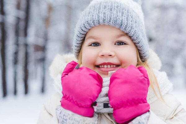 close-up portrait of cute little child in hat and mittens smiling at camera in winter park