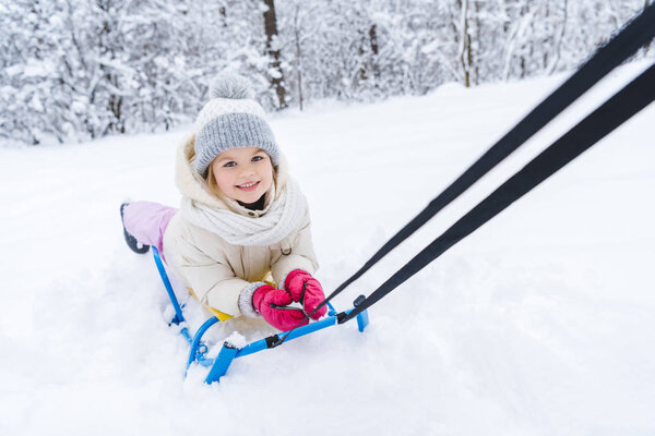 cute little child lying on sled and smiling at camera in winter park