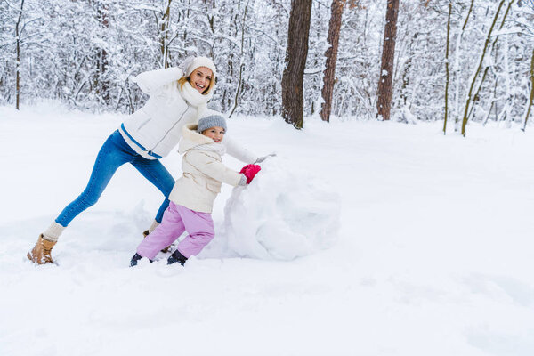 happy mother and daughter making snowman and smiling at camera in winter park