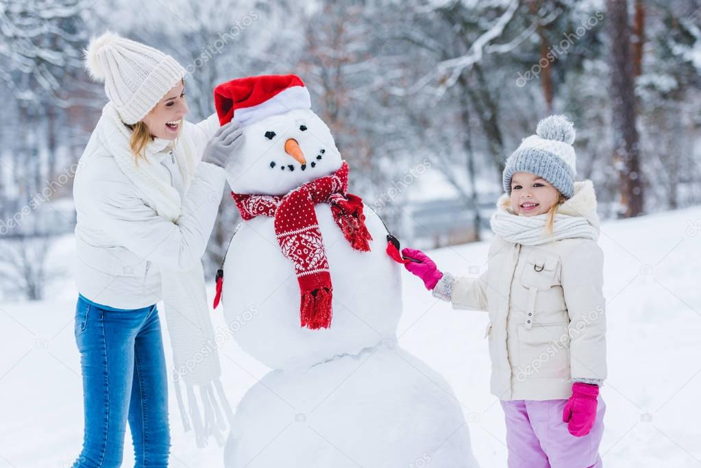 happy mother and daughter making snowman together in winter forest
